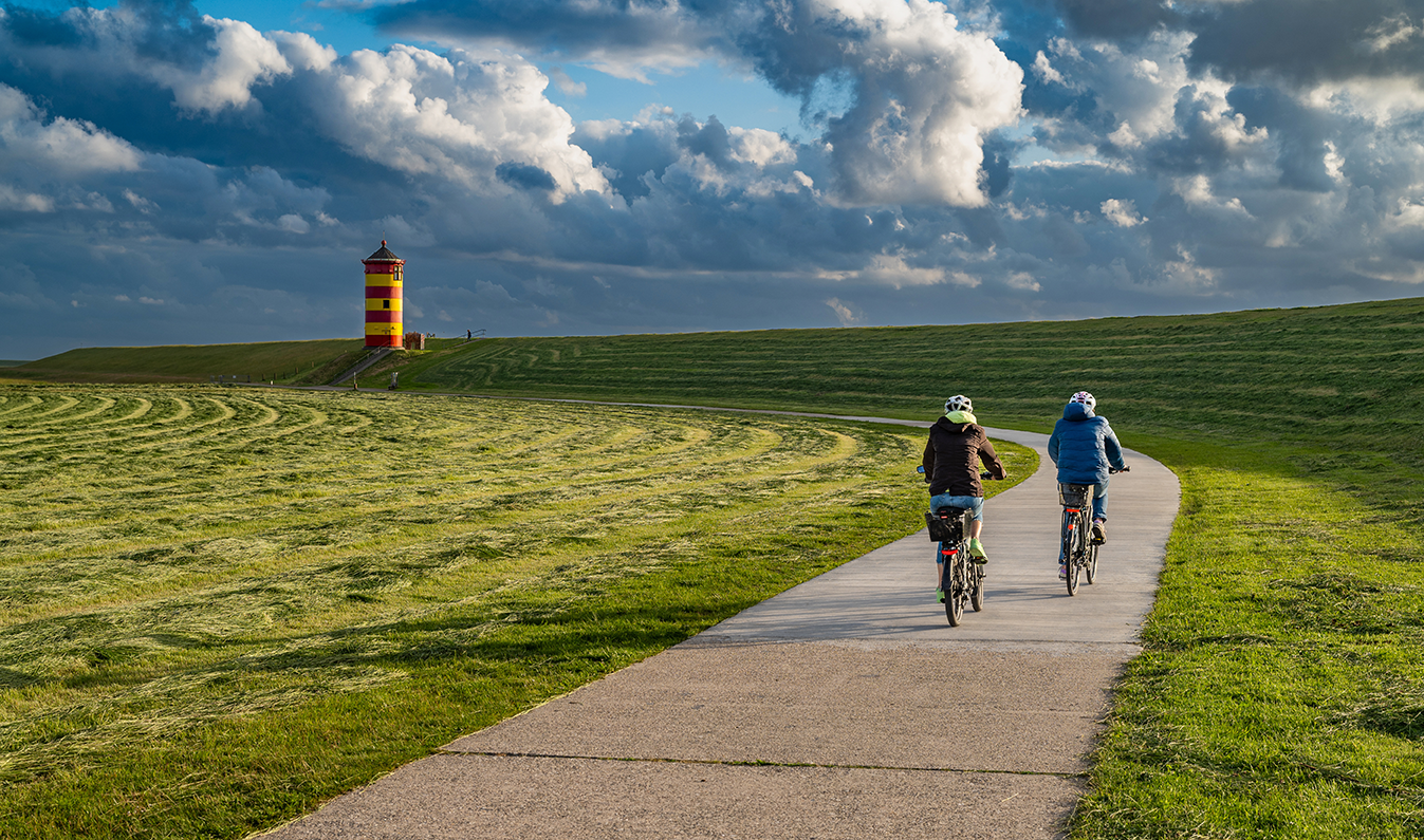 Zwei Radfahrer genießen die Landschaft in Greetsiel, mit dem markanten Leuchtturm im Hintergrund.
