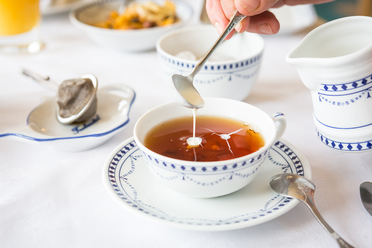 Eine Hand gießt Milch in eine Tasse Tee, umgeben von eleganten Tellern, typisch für eine Tee - Zeremonie im Romantik-Hof Greetsiel.