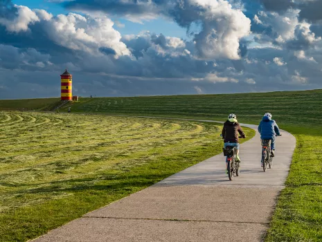 Zwei Radfahrer genießen die Landschaft in Greetsiel, mit dem markanten Leuchtturm im Hintergrund.