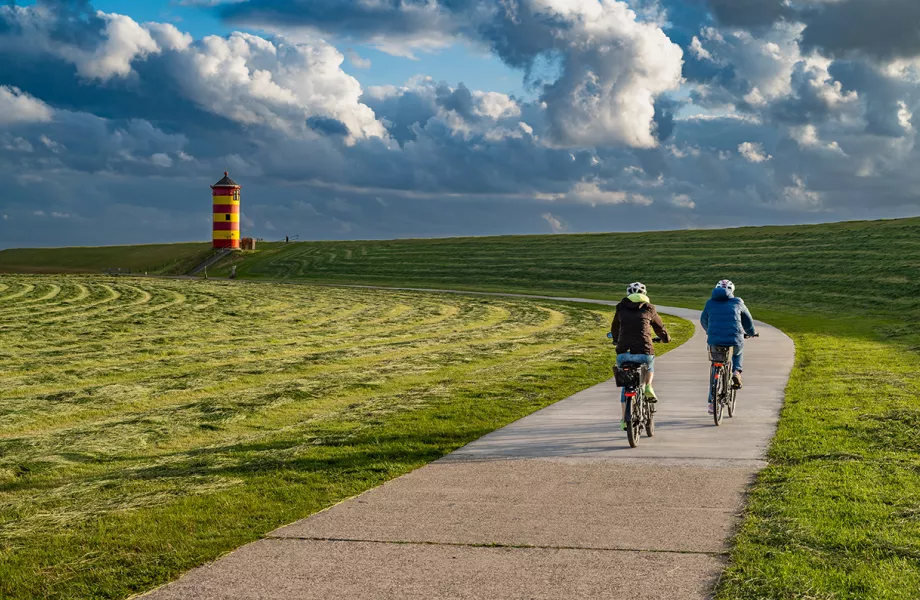 Zwei Radfahrer genießen die Landschaft in Greetsiel, mit dem markanten Leuchtturm im Hintergrund.
