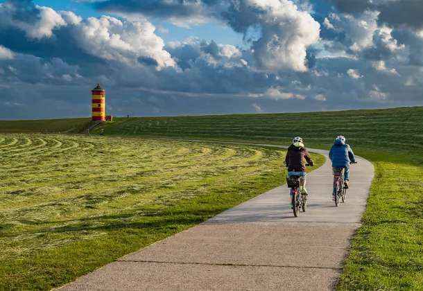 Zwei Radfahrer genießen die Landschaft in Greetsiel, mit dem markanten Leuchtturm im Hintergrund.